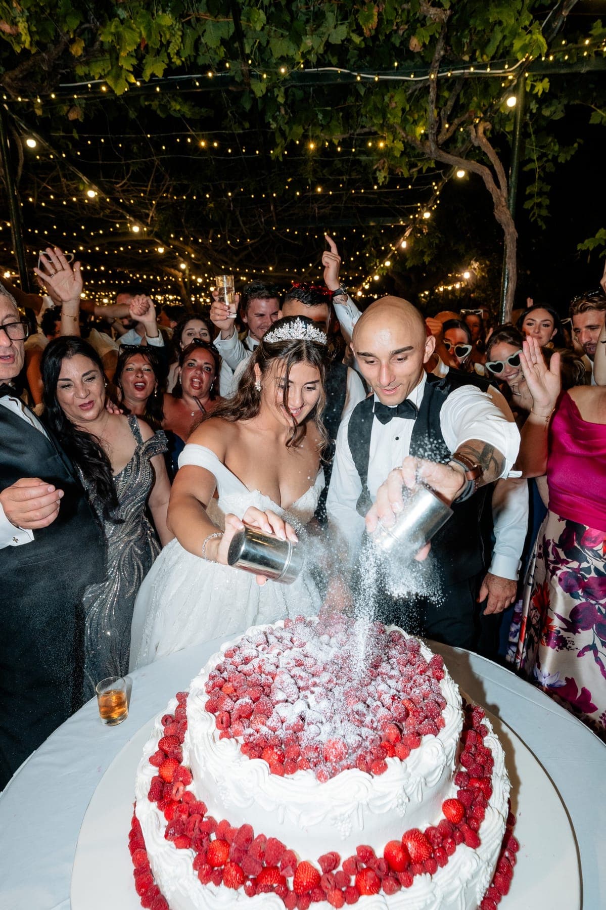 Bride and groom dusting their strawberry wedding cake as guests celebrate under string lights