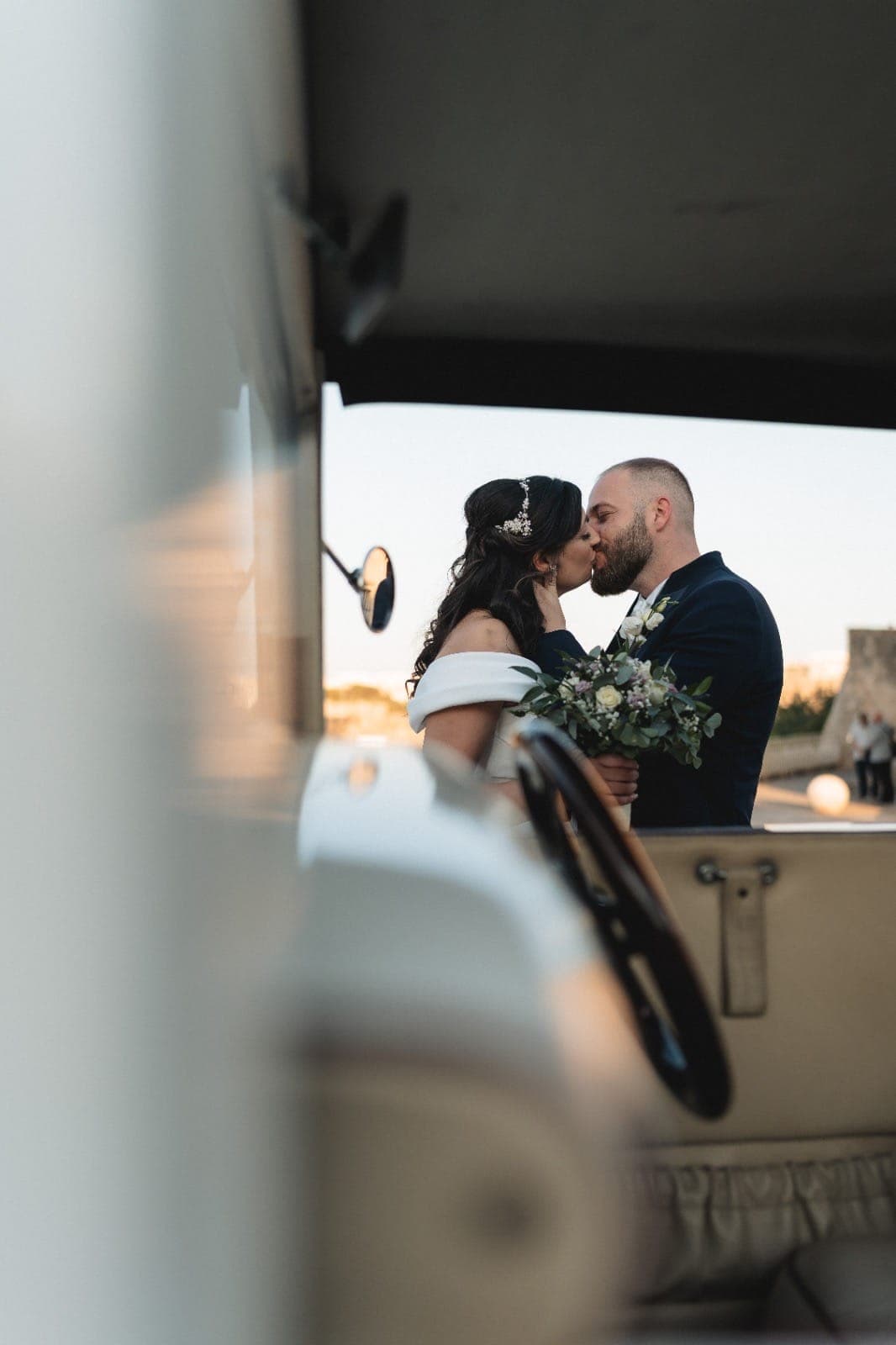 Newlyweds sharing a kiss framed through the window of a vintage wedding car at sunset